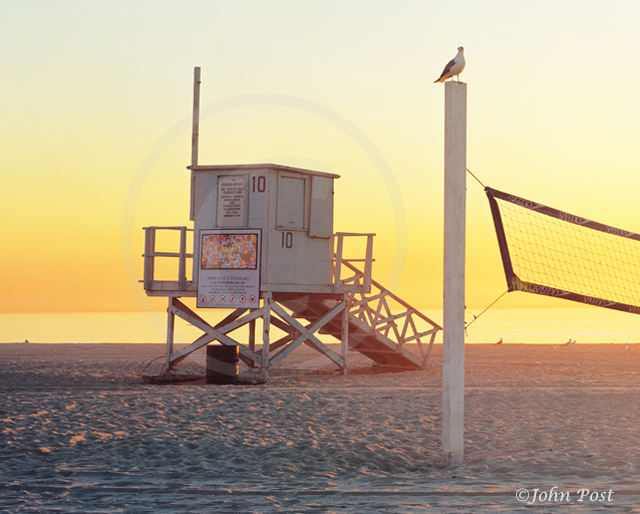 Lifeguard tower on beach, clear sunset sky with volleyball net and seagull on top of pole.
